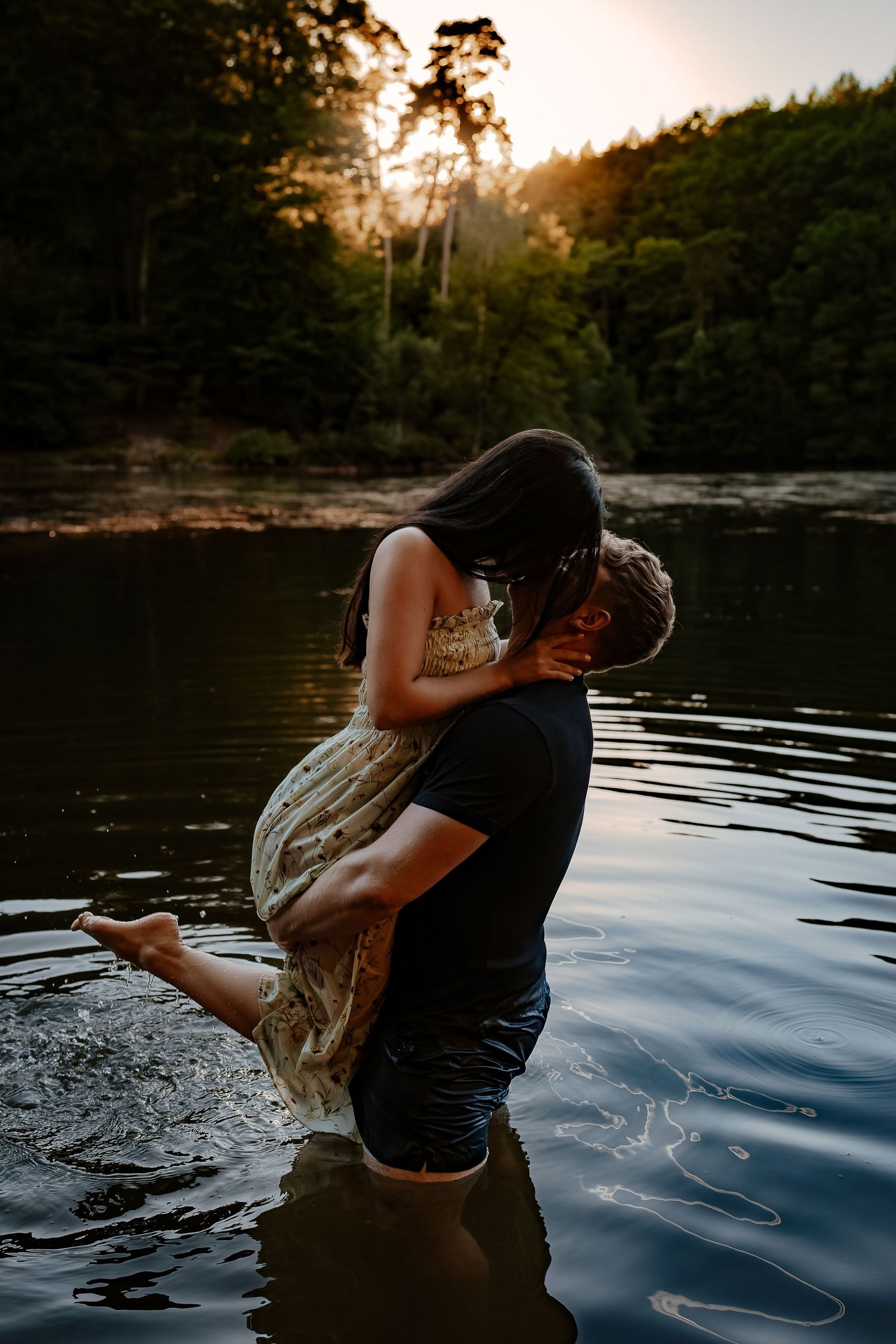 Romantic couple embracing in lake water at sunset with forest backdrop.