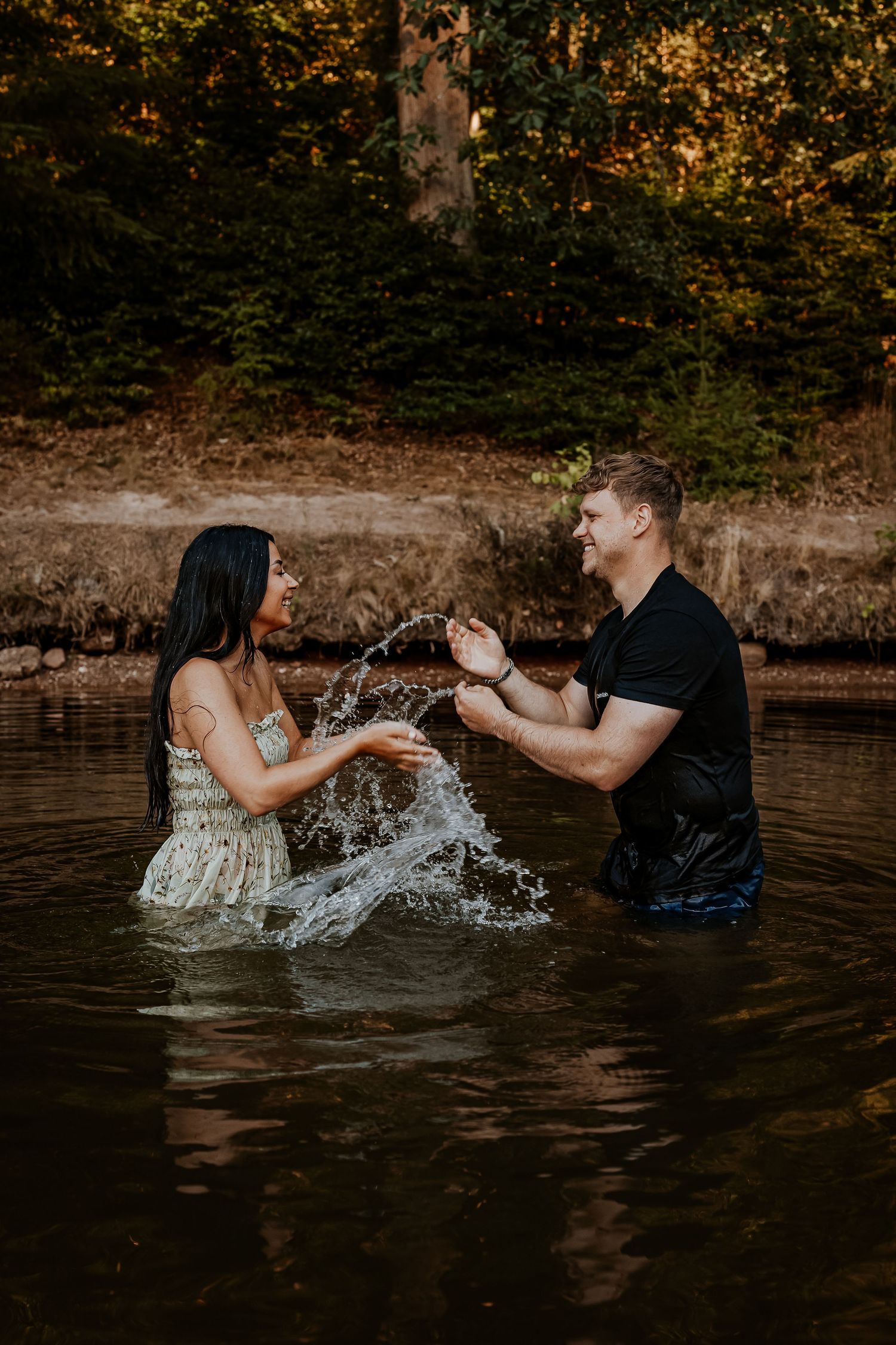 Playful couple splashing water at each other in lake during sunset.