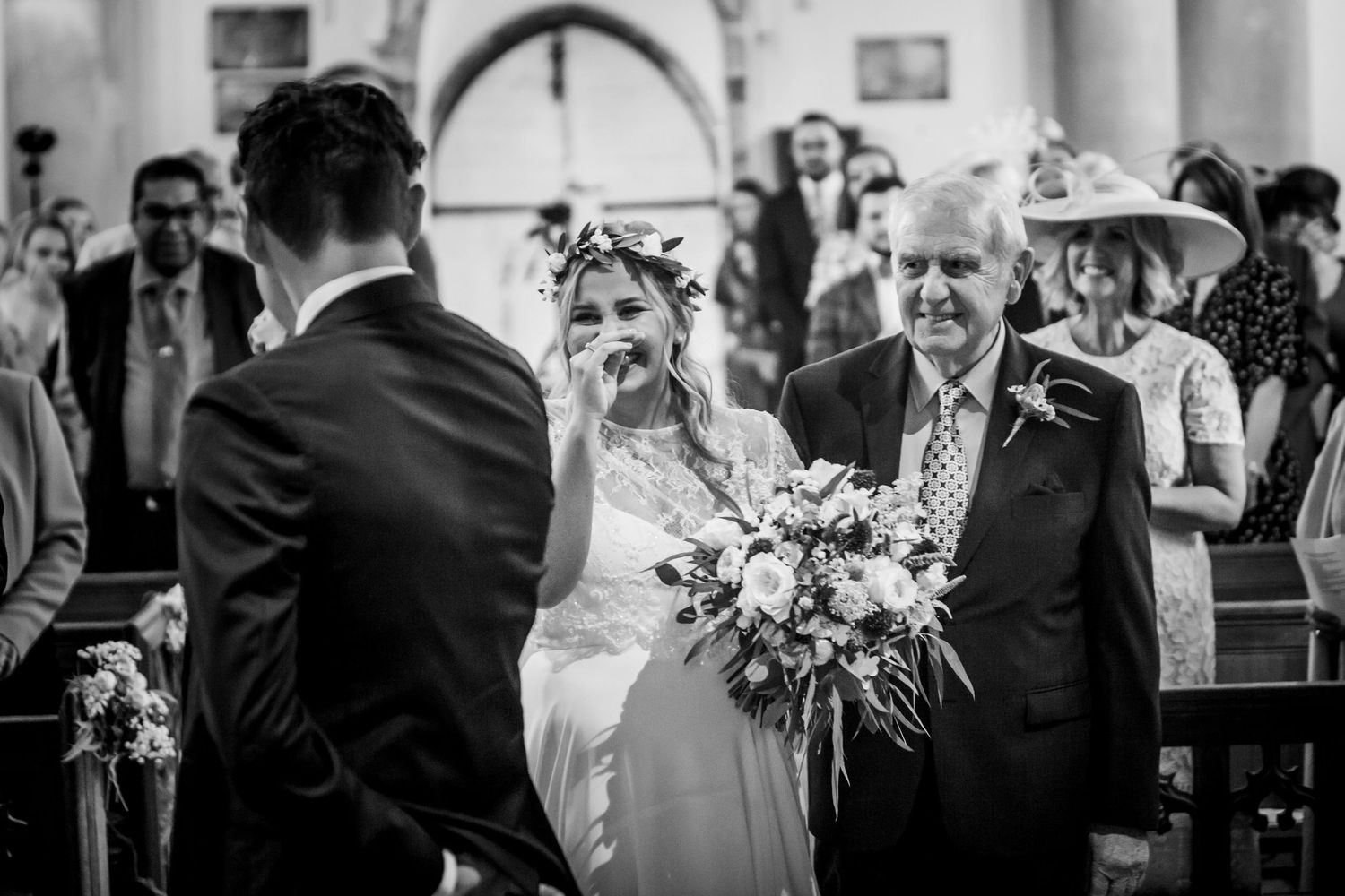 Bride walks down church aisle with her father, holding a bouquet, in a black and white wedding ceremony photo.