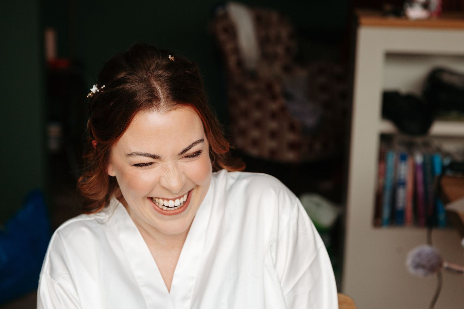 Bride laughing joyfully in white robe with hair accessories during wedding morning preparations.