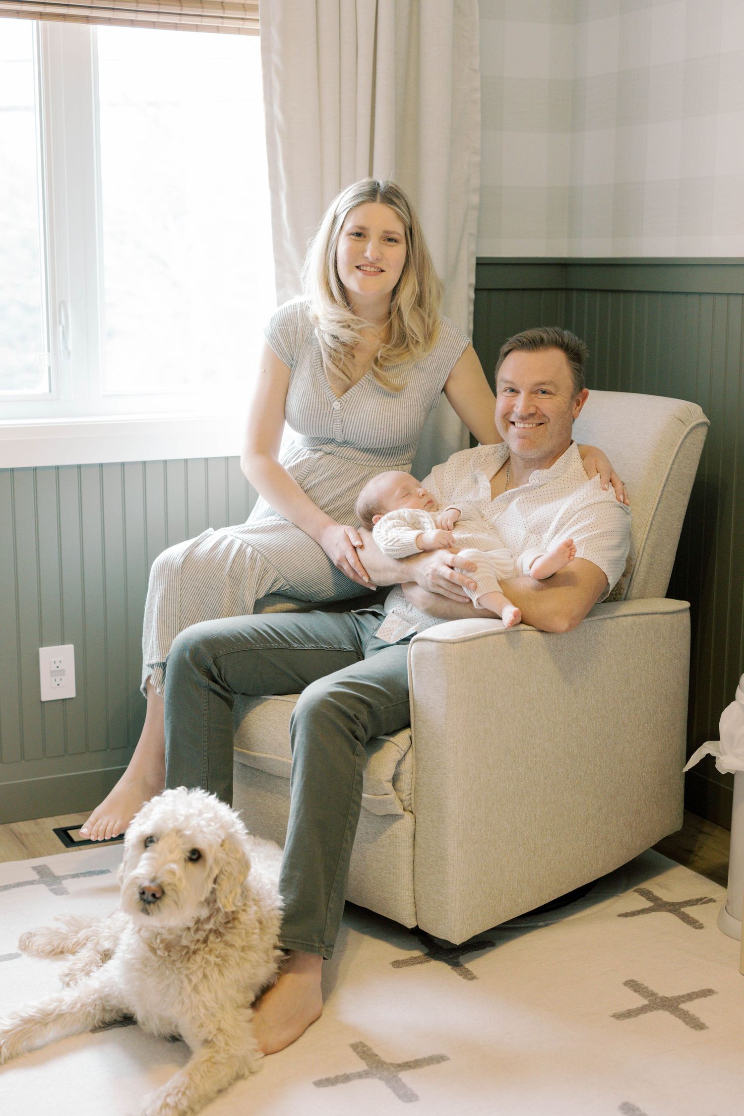A couple sits in a nursery chair with their newborn baby while a white fluffy dog rests on the floor nearby.