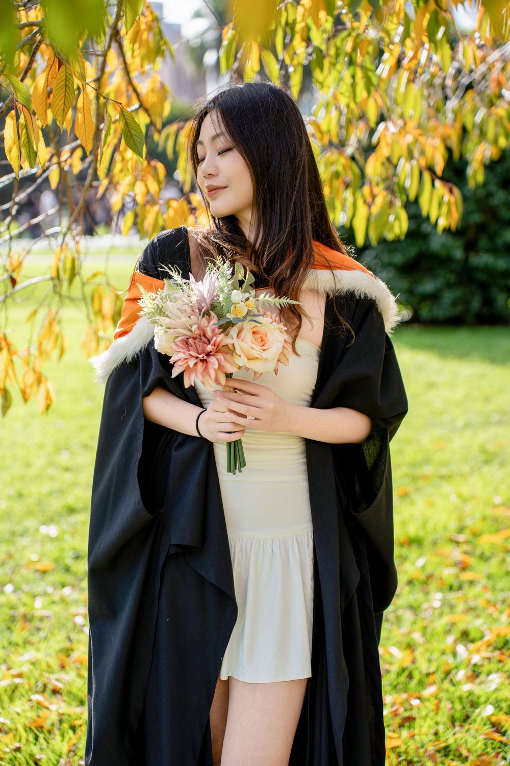 A graduation portrait of a student holding a bouquet in a scenic outdoor setting in Auckland.