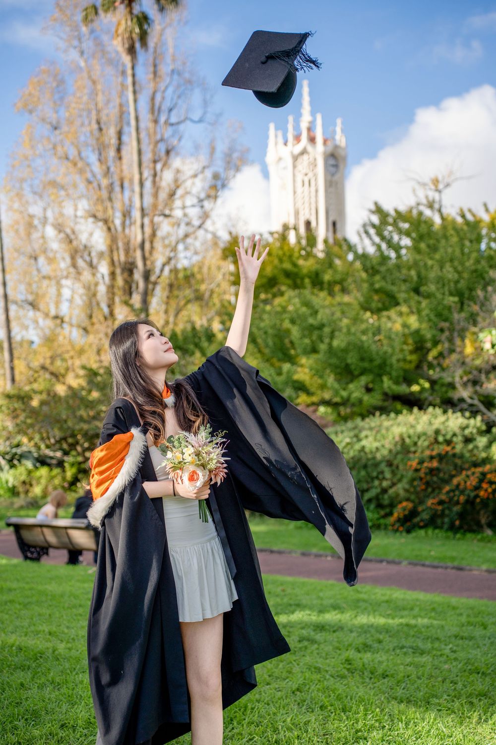 A joyful graduate throwing her cap into the air in a scenic Auckland park.