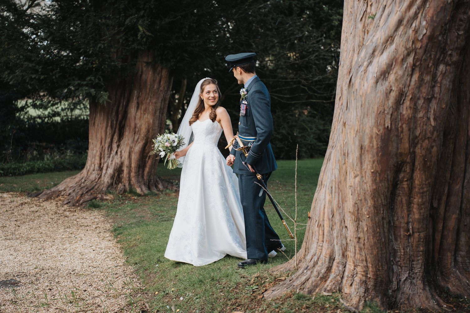 Bride in white gown and groom in military uniform share a romantic moment among ancient trees on their wedding day at Caswell House