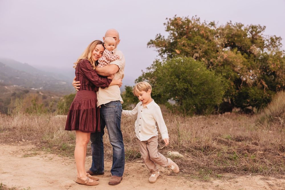 Family of four posing outdoors on a dirt path with misty hills and green trees in the background.