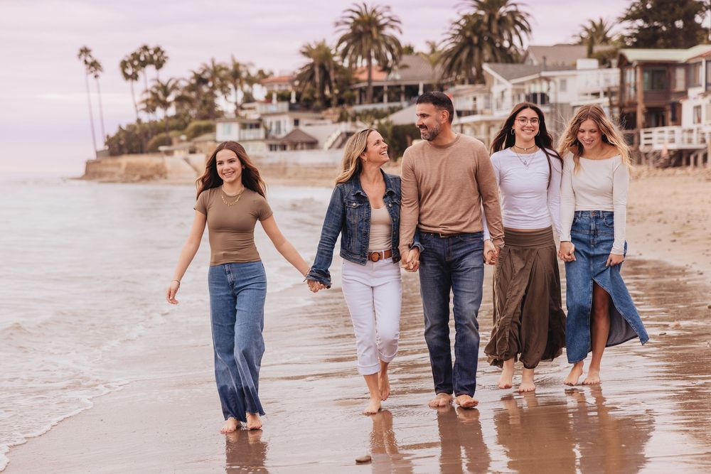 Family of five walking barefoot on a sandy beach near Malibu homes, holding hands and smiling together.
