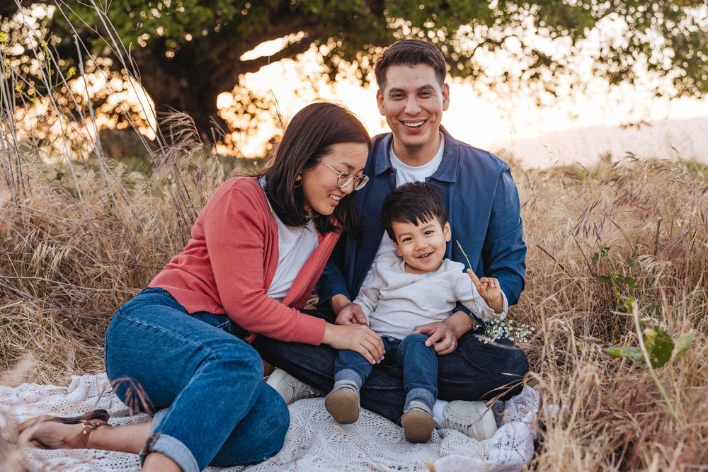 Family of three sitting on blanket in golden field at sunset, parents smiling at toddler boy.