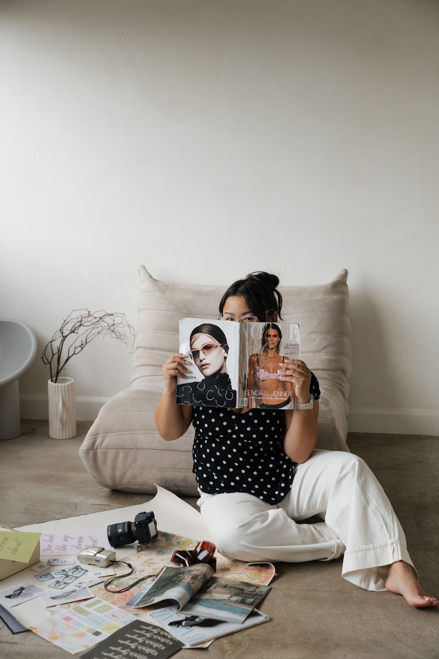 Woman sitting on floor holding fashion magazines near a beige sofa, surrounded by photos, a camera, and papers.