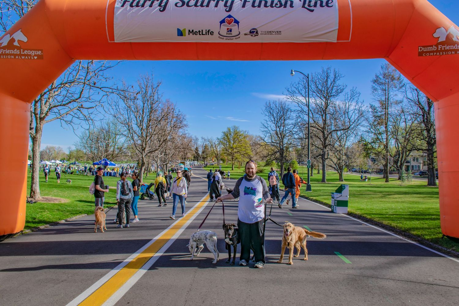 Person with two dogs at Furry Scurry Finish Line charity race event in a sunny park with orange arch.