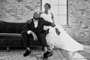 Black and white wedding photo of bride and groom posing on a velvet sofa in a rustic brick-walled venue.