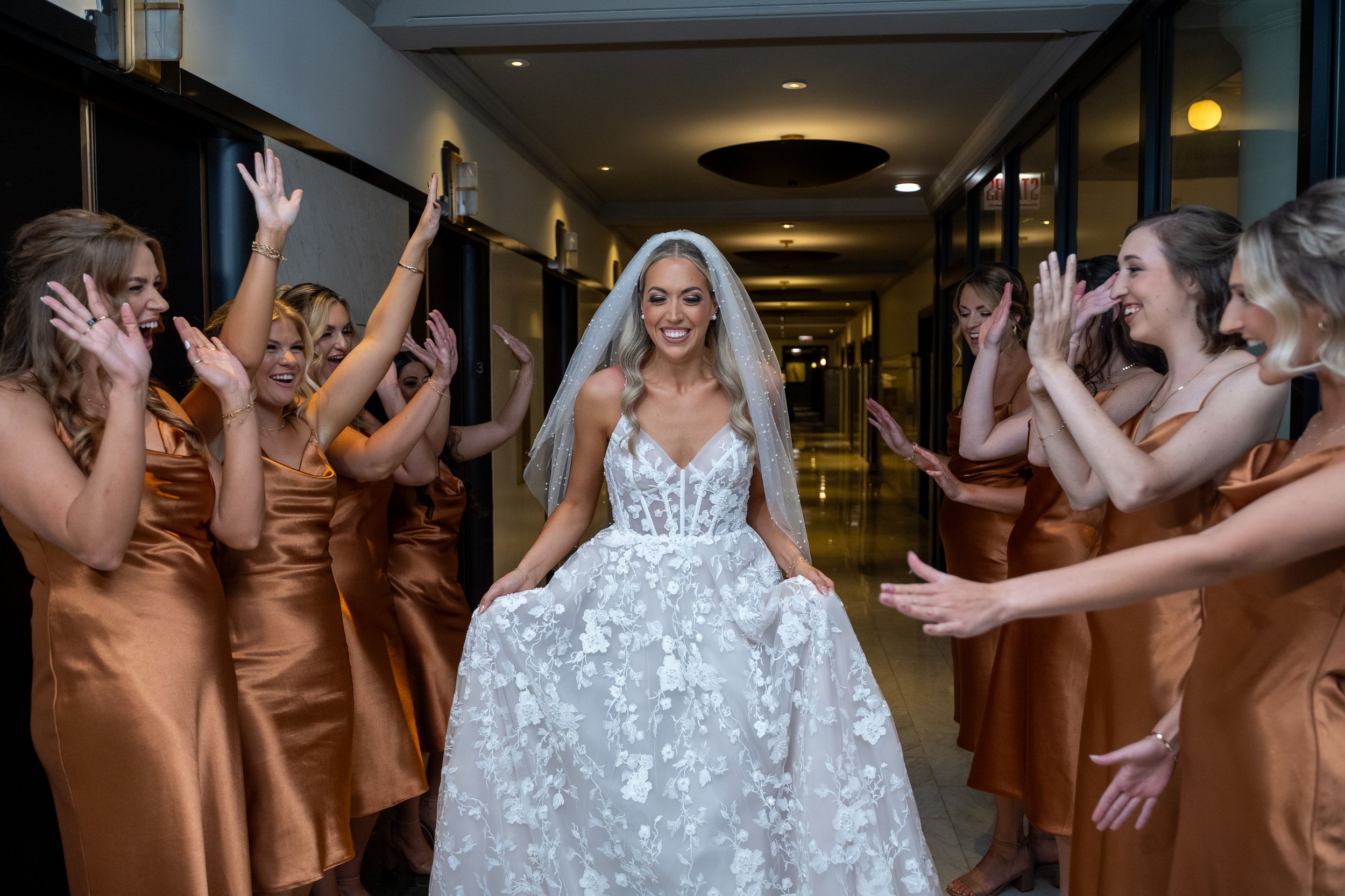 Bride in floral ballgown walks down hotel hallway while bridesmaids in copper satin dresses cheer around her.