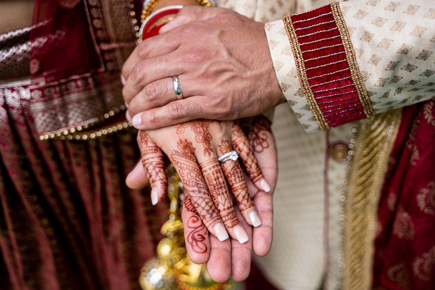 Close-up of Indian bride and groom hands with mehndi henna, wedding rings, and traditional red and gold attire.