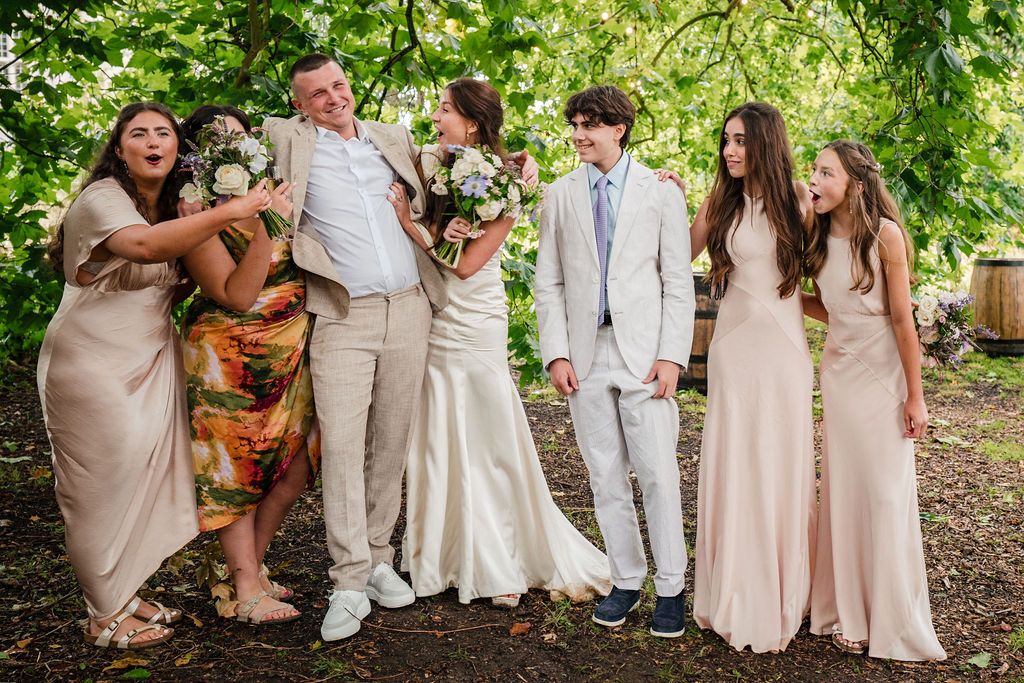Wedding party laughing outdoors, bride in white gown with bouquet, groom in beige suit, bridesmaids in blush pink dresses.