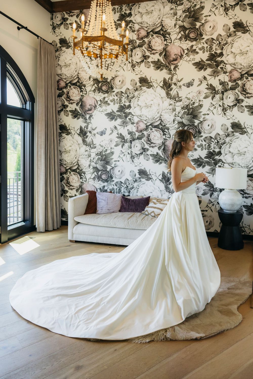 Bride in elegant white gown with long train stands in room with floral wallpaper and chandelier getting ready for her wedding in  Wisconsin