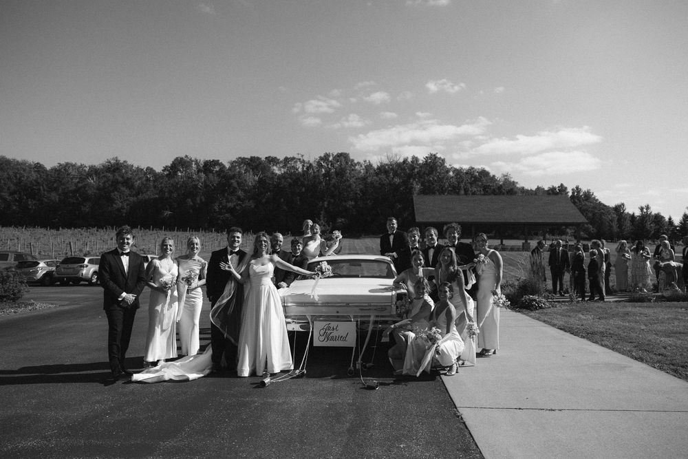 Black and white wedding party photo with bride, groom, and guests posing beside a classic car outdoors in  Wisconsin