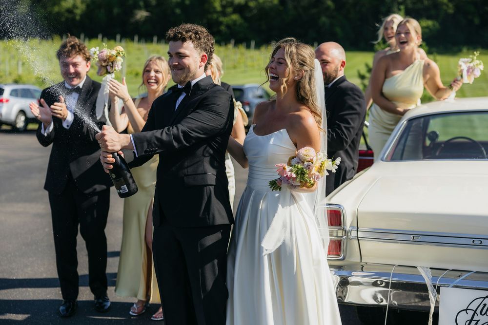 Bride and groom popping champagne bottle outside classic white car with wedding party celebrating in background in  Wisconsin