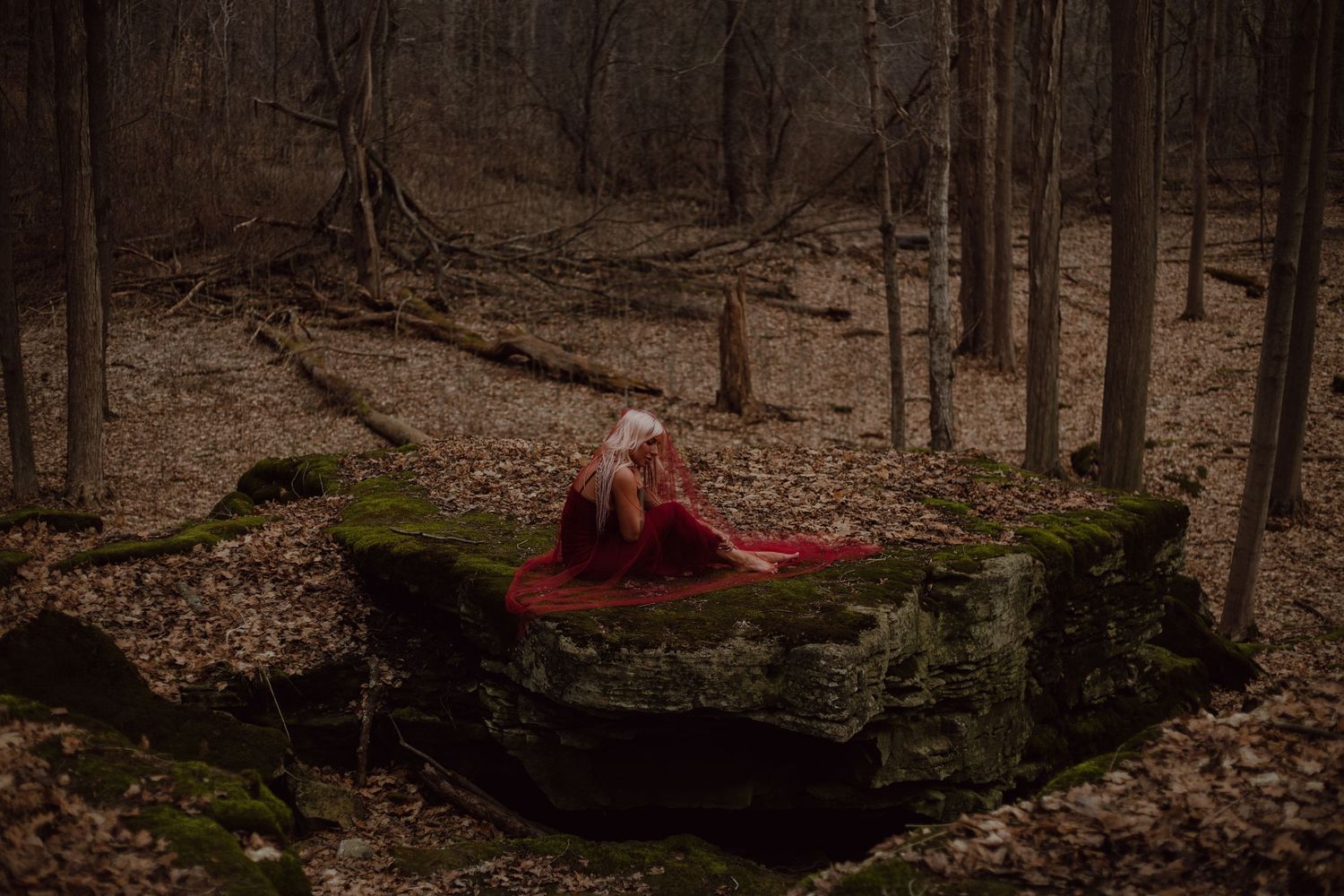 Model crouched on a mossy rock while red fabric drapes over her in the forest to make this session dramatic and moody.