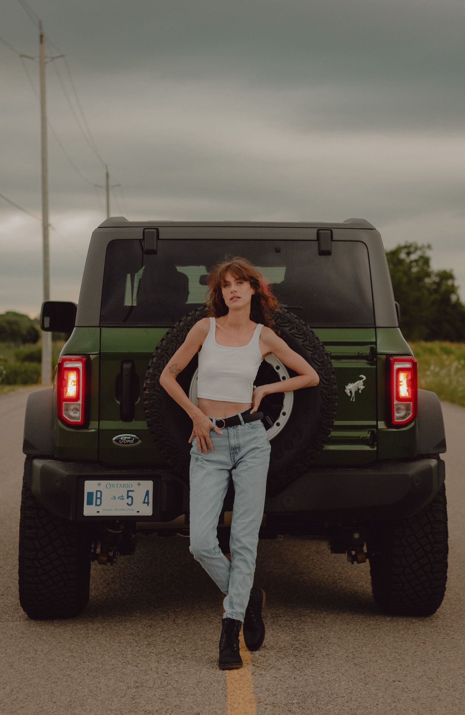 Model posing on the road in front of green Bronco. Model is wearing blue jeans and a white tank top.