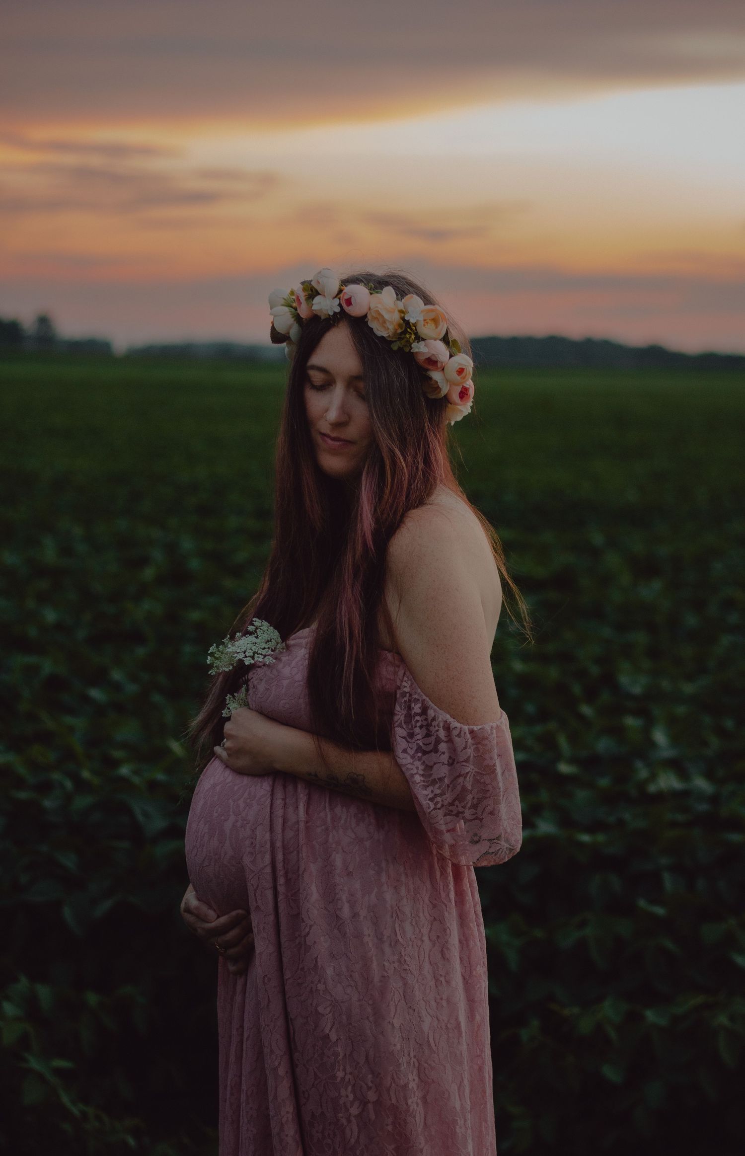Maternity session in farmer's field right after sunset, during blue hour. Mother to be is wearing a flower crown and flowing pink dress.