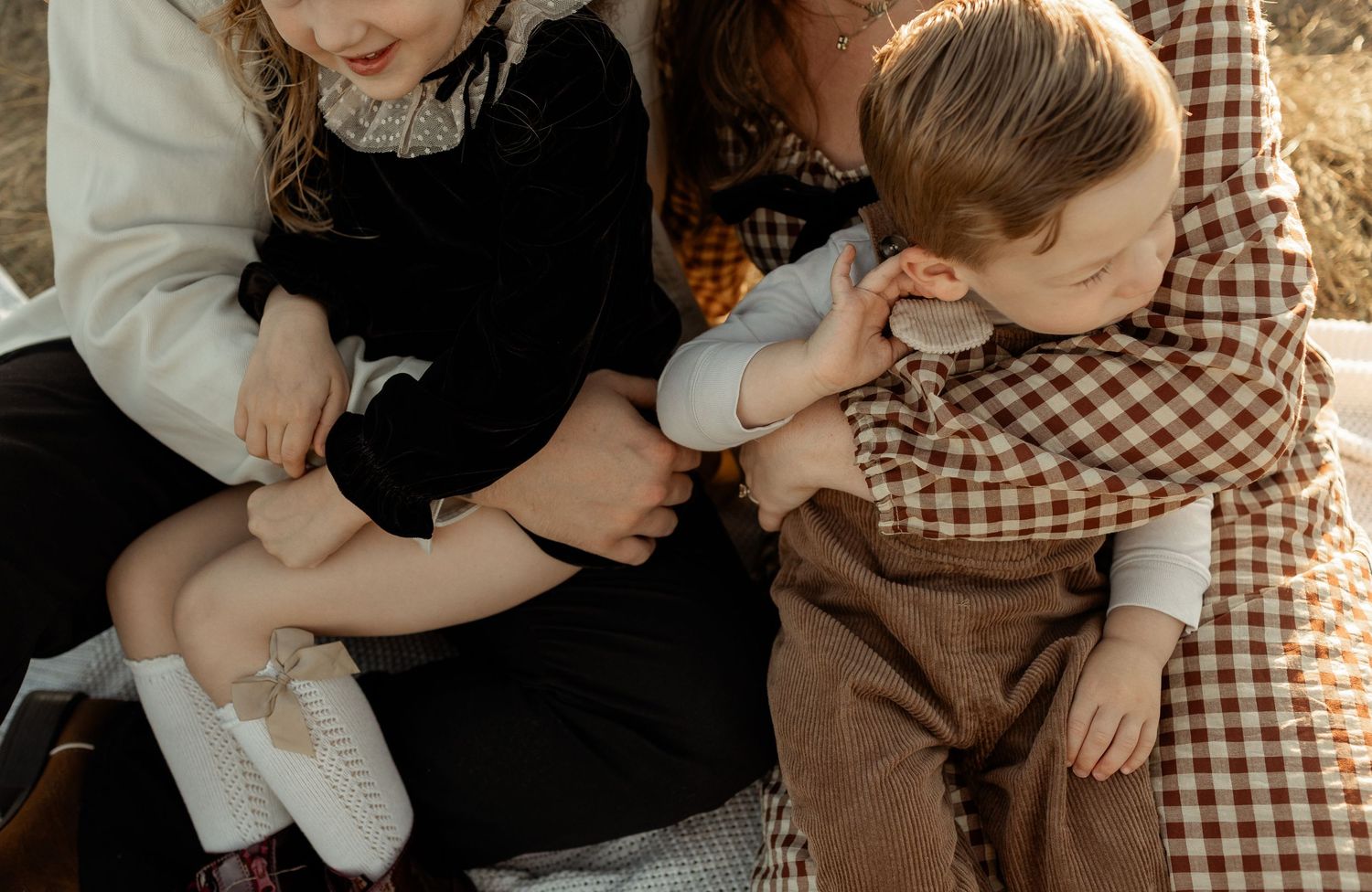 Two children and an adult sit together outdoors, with a toddler in plaid drinking from a bottle.