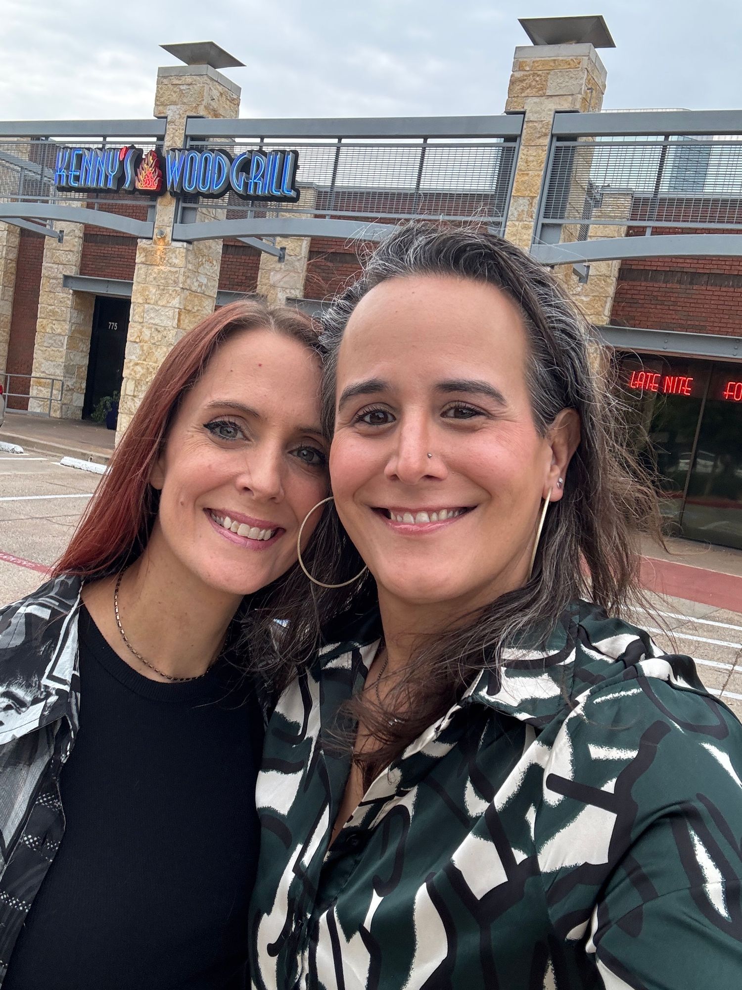 Two smiling women taking a selfie outside Henry's Wood Grill restaurant on a cloudy day.