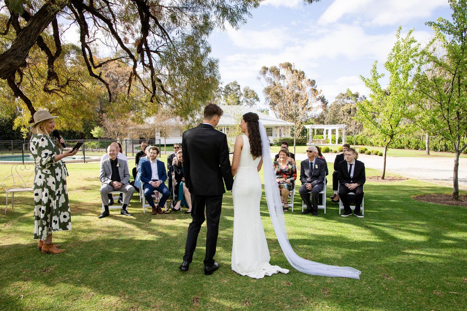 Outdoor wedding ceremony on green lawn with bride and groom facing officiant, guests seated in chairs under trees.