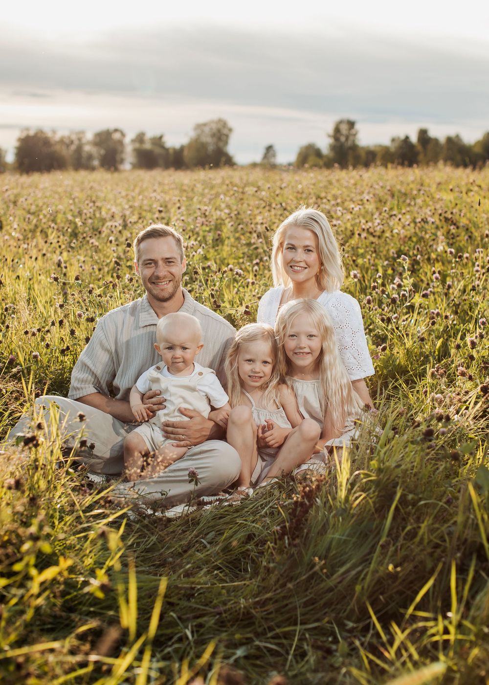 Family of five posing in a sunny wildflower field, parents smiling with three young children during golden hour.