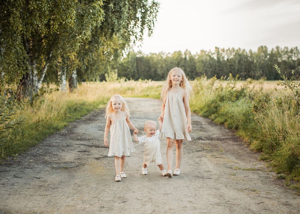 familjefotografering på sommaren på en skogsväg i Boden med tre syskon i matchande beiga färger som går hand i hand.