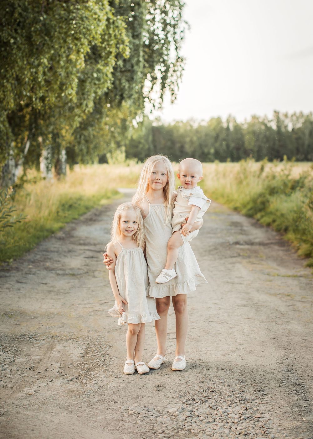familjefotografering på sommaren på en skogsväg i Boden med tre syskon i matchande beiga färger som kramar om varandra.