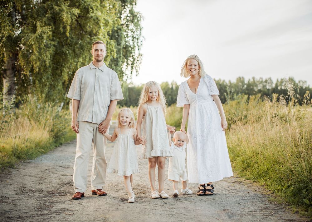 Happy family of five in white outfits walking on a sunny dirt path surrounded by green fields and trees.