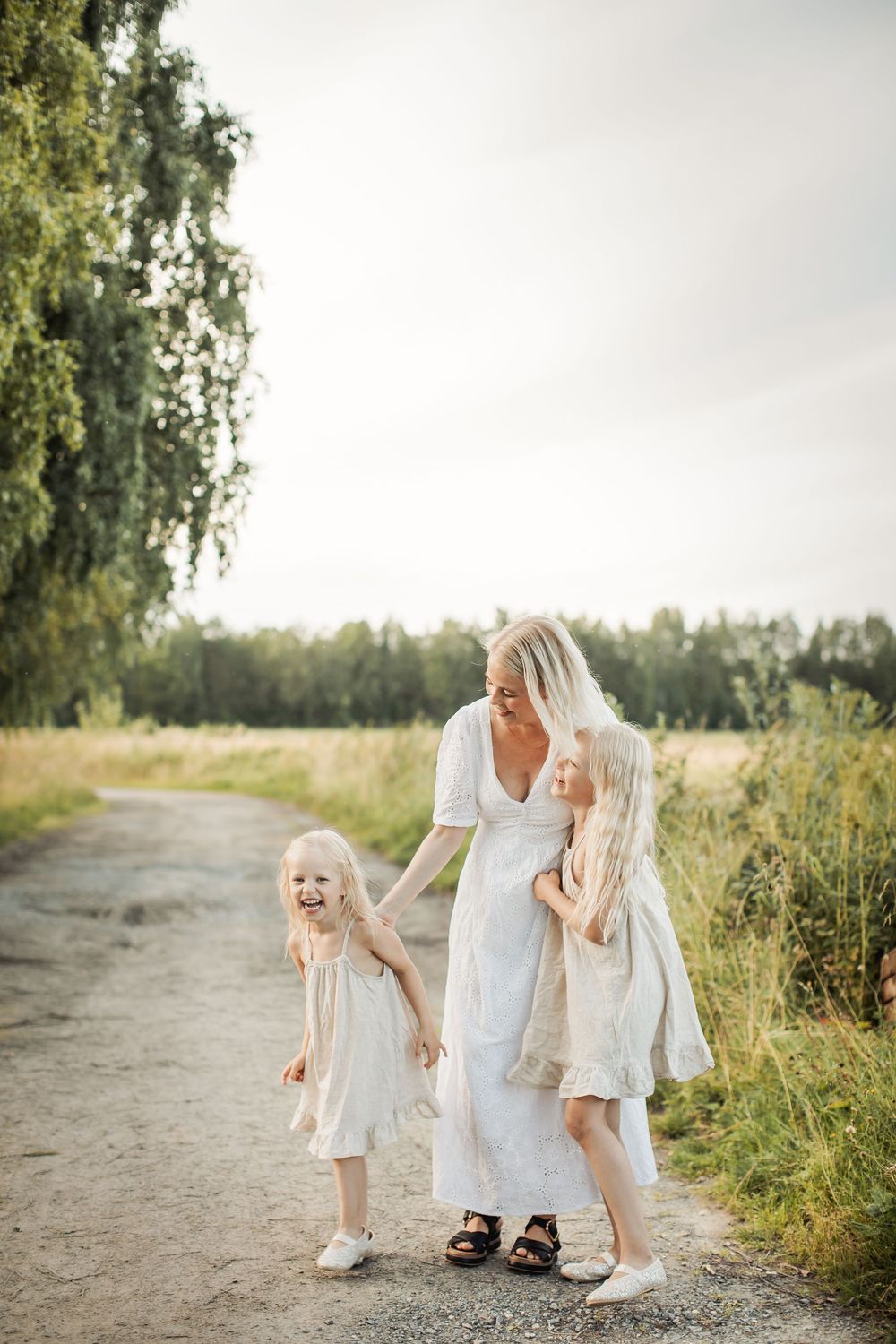 A blonde mother in a white dress walks with her two young daughters in matching white dresses on a rural path.