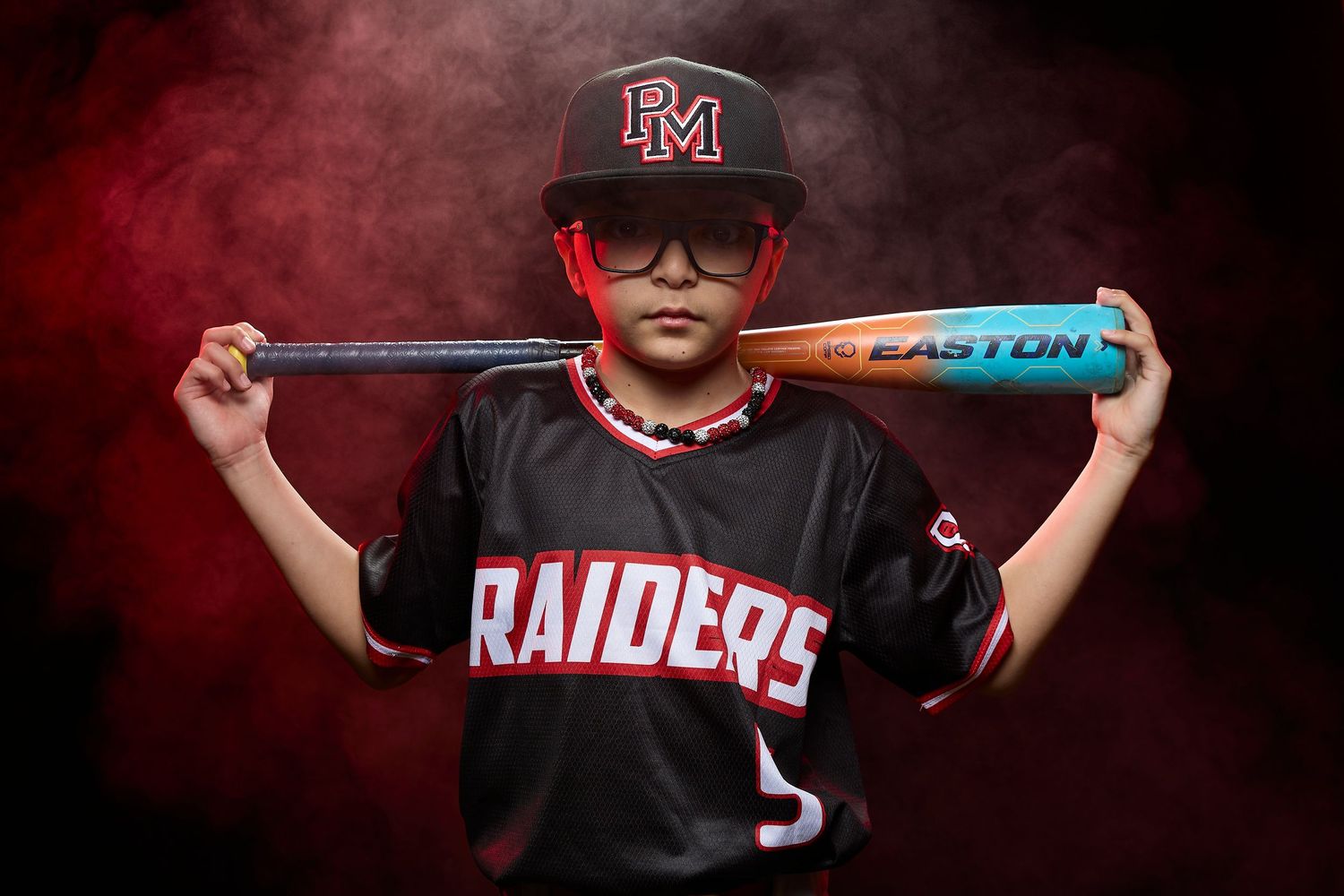 Youth baseball player in Raiders uniform poses with blue bat against dramatic red smoky background.