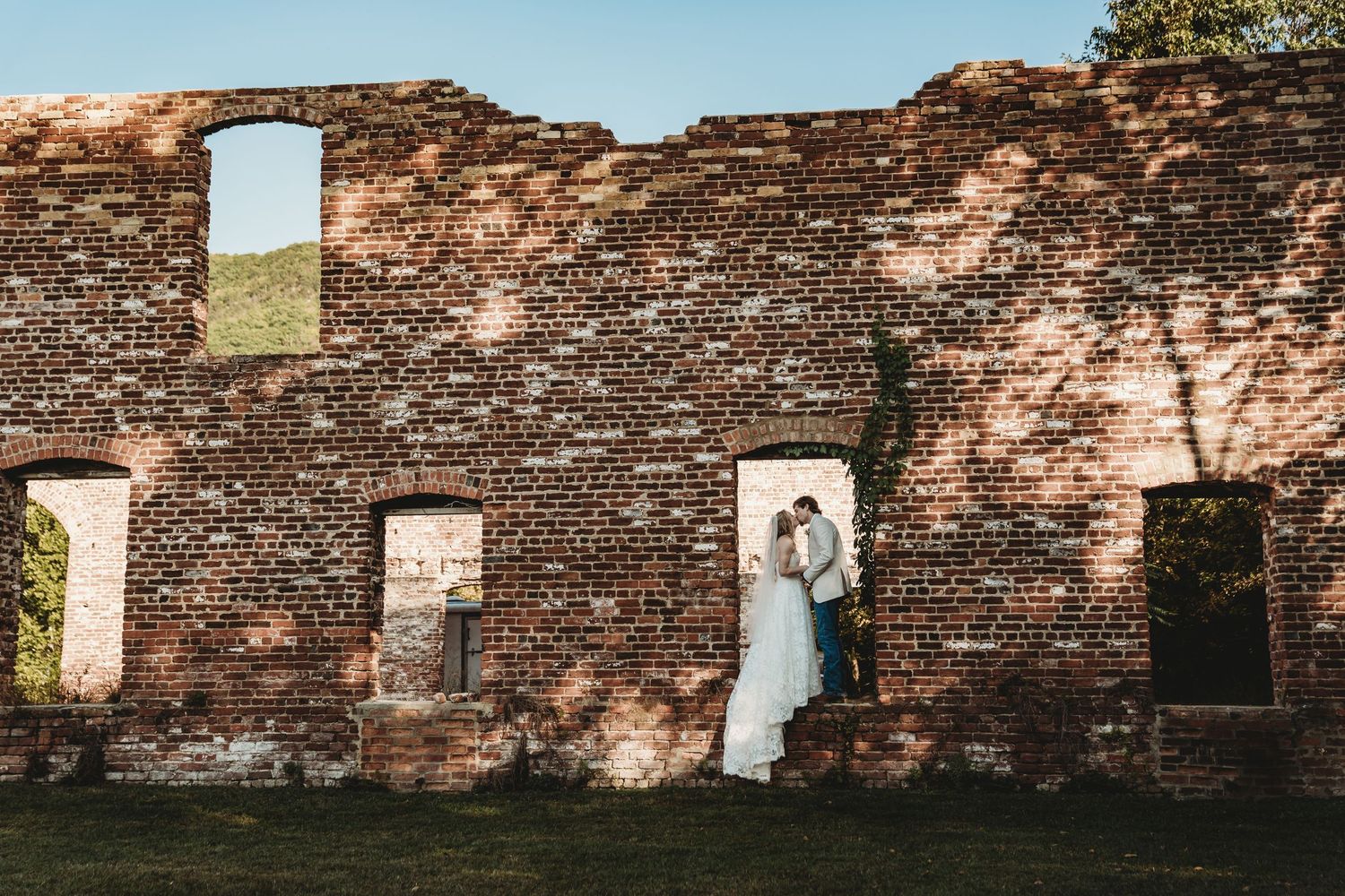 Wedding party celebrates with joyful dip kiss pose in front of stone mansion on grassy lawn.