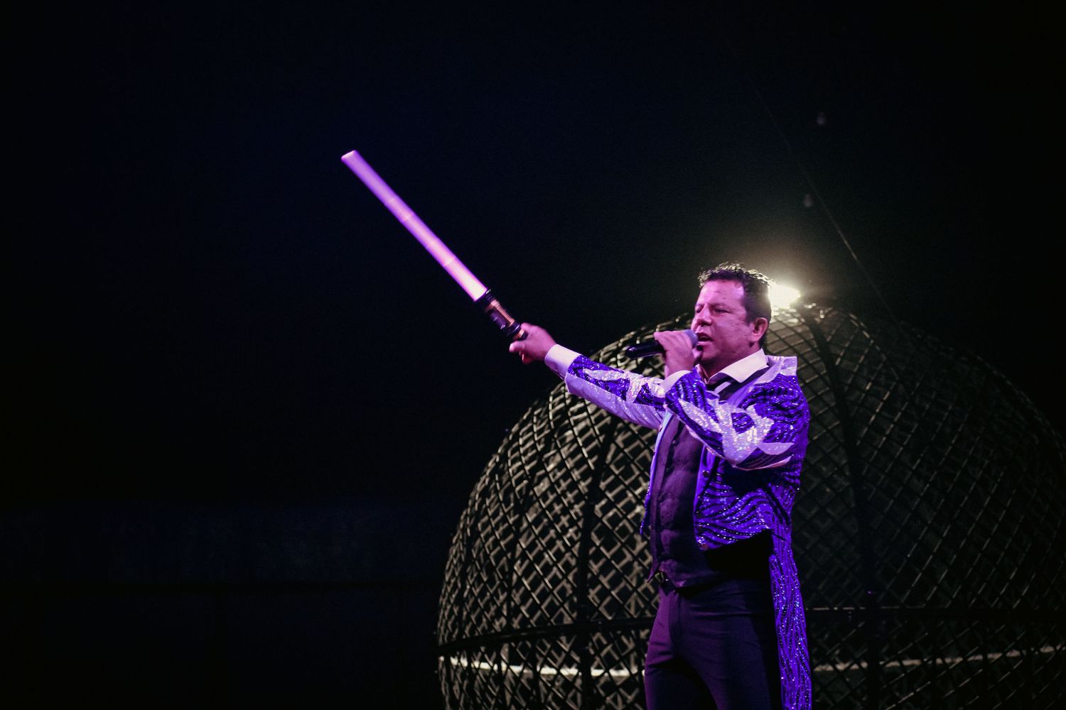 Performer on stage points upward during a concert with dramatic purple lighting and fog effects in the background.