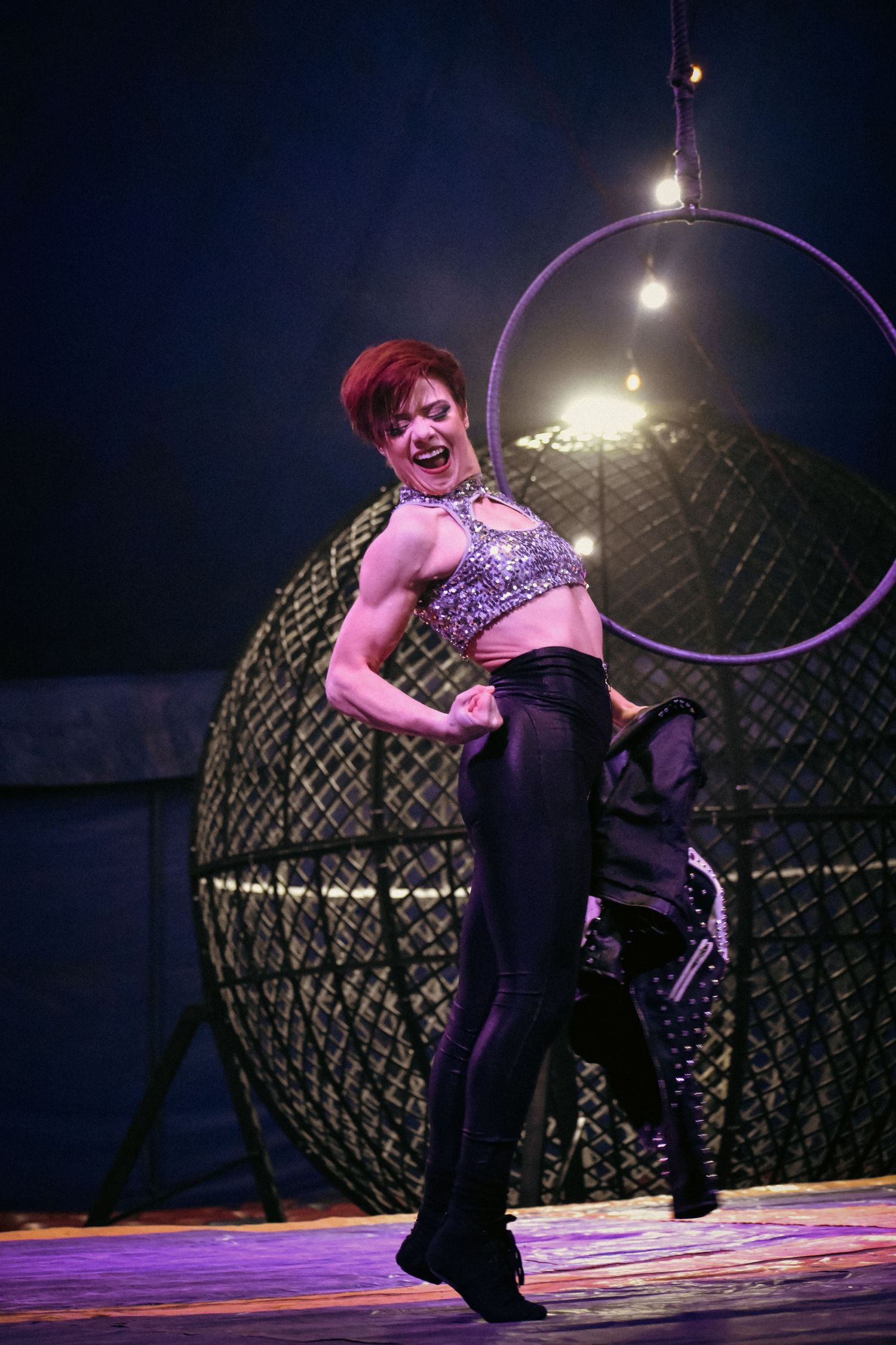 Circus performer in sparkly costume spins with hula hoop on stage during dramatic nighttime performance.