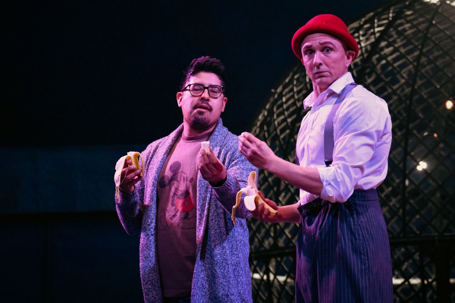 Two performers interact on stage during a theatrical performance, one wearing a cardigan and one in a dress shirt and cap.
