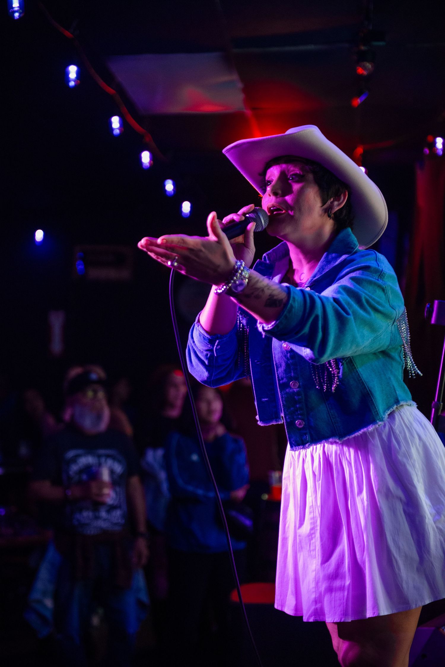 Performer in a white dress and hat sings passionately on stage under dramatic purple and blue lighting at a concert venue.