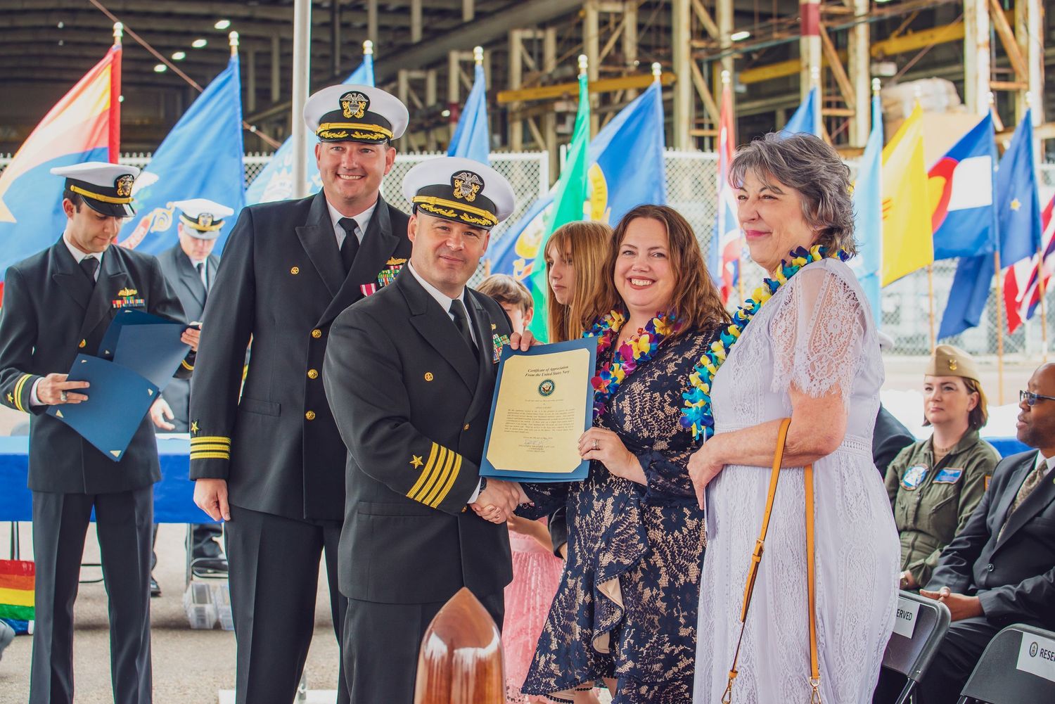 Military officers and attendees gather for an award ceremony with colorful flags displayed in the background.