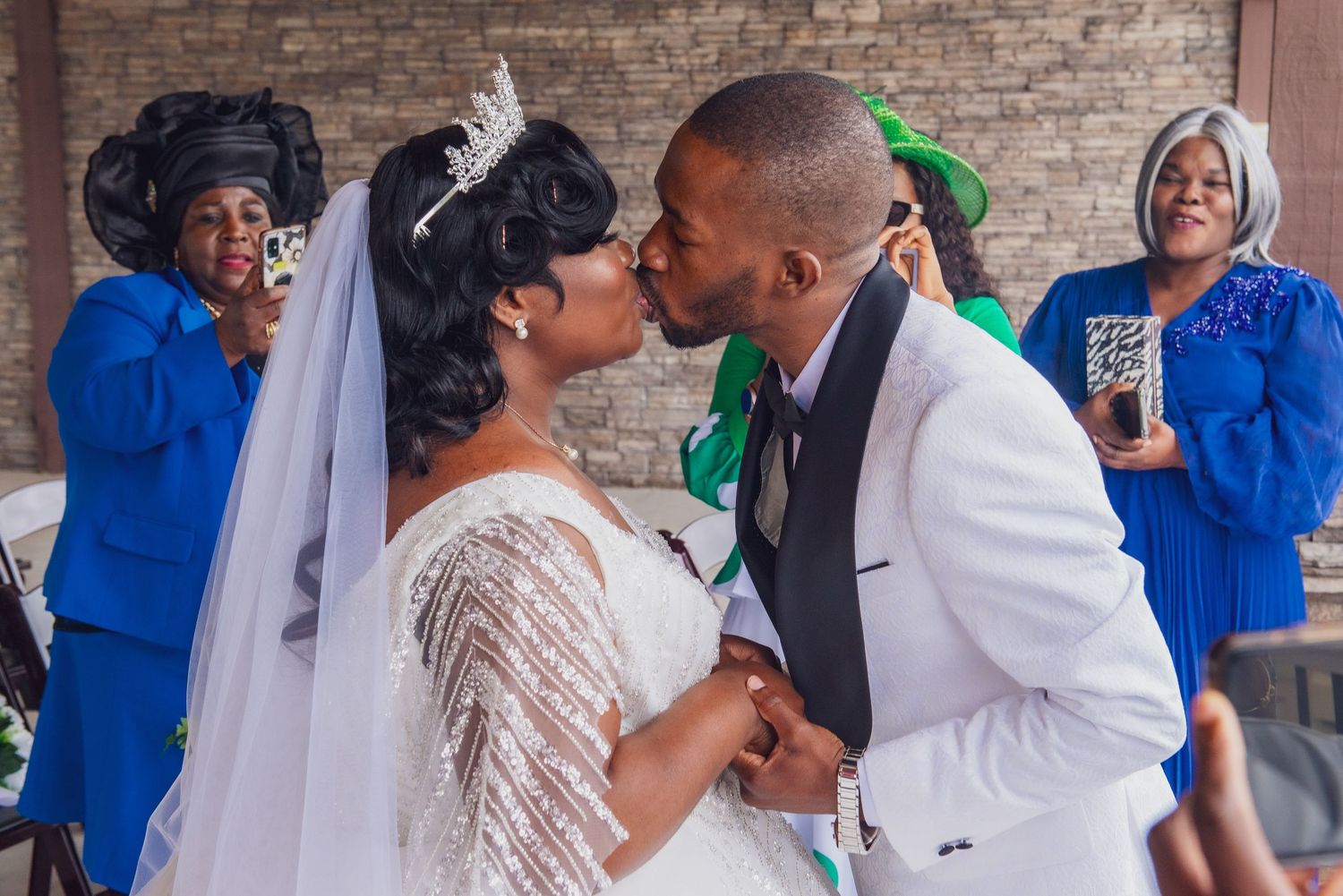 A couple shares their first kiss as newlyweds at their wedding ceremony while guests in blue and green look on.