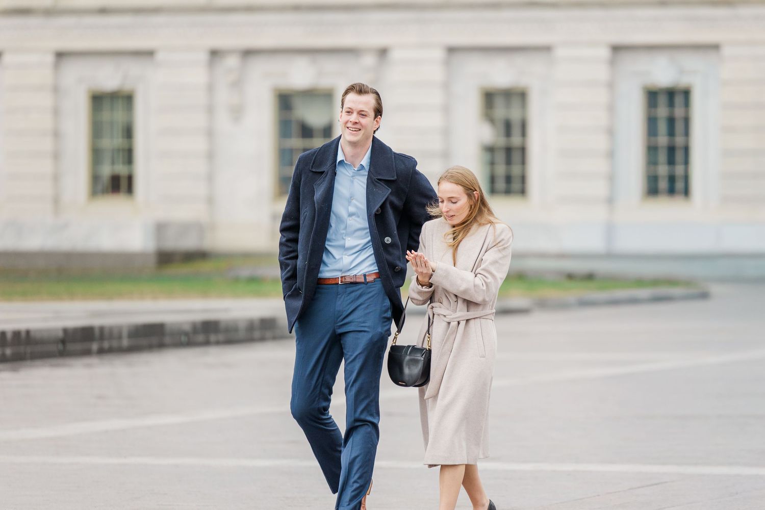 A couple in formal attire walks together on a sunny day outside a grand stone building.