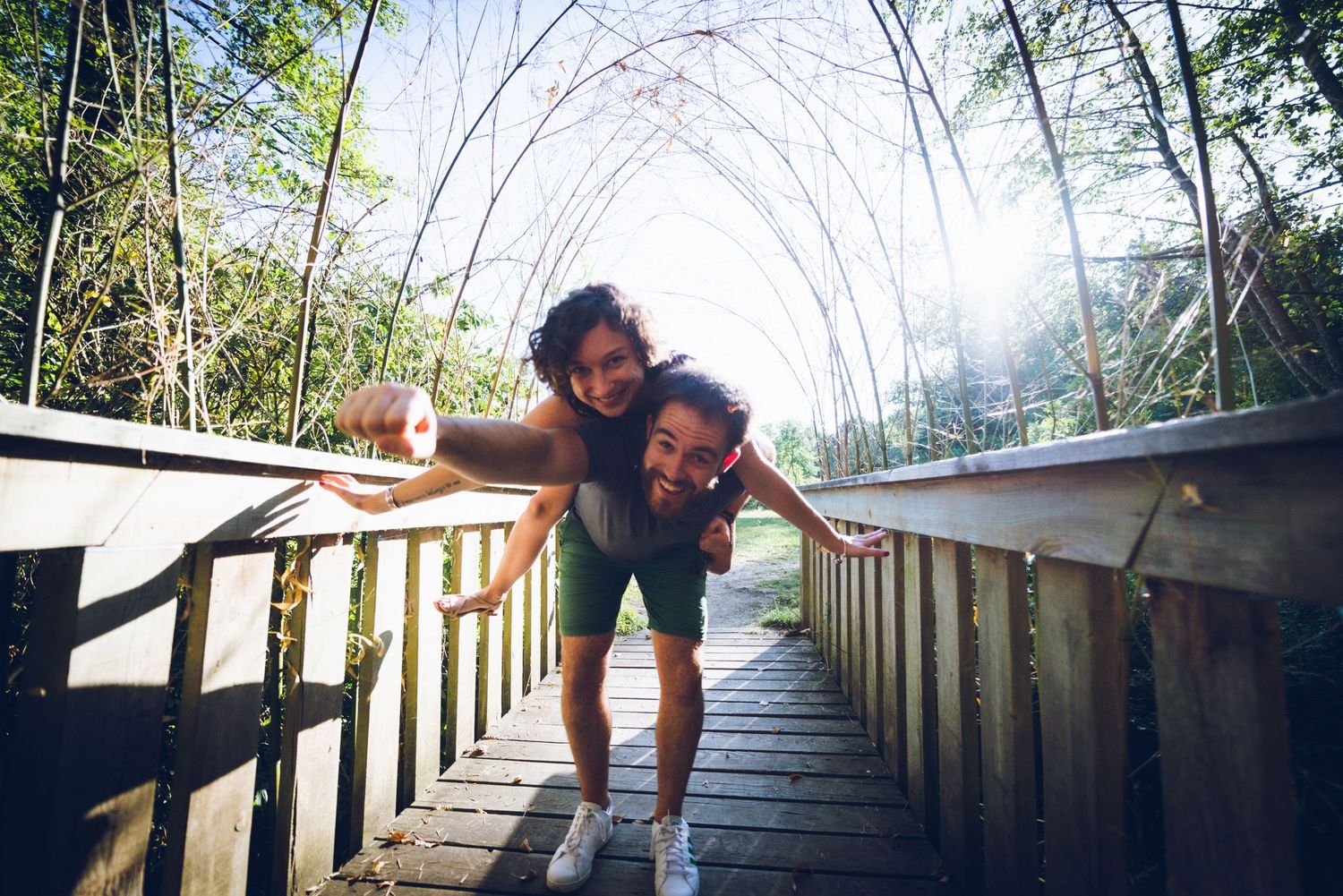 Couple souriant marchant sur une passerelle en bois entourée de végétation lors d’une séance photo engagement.