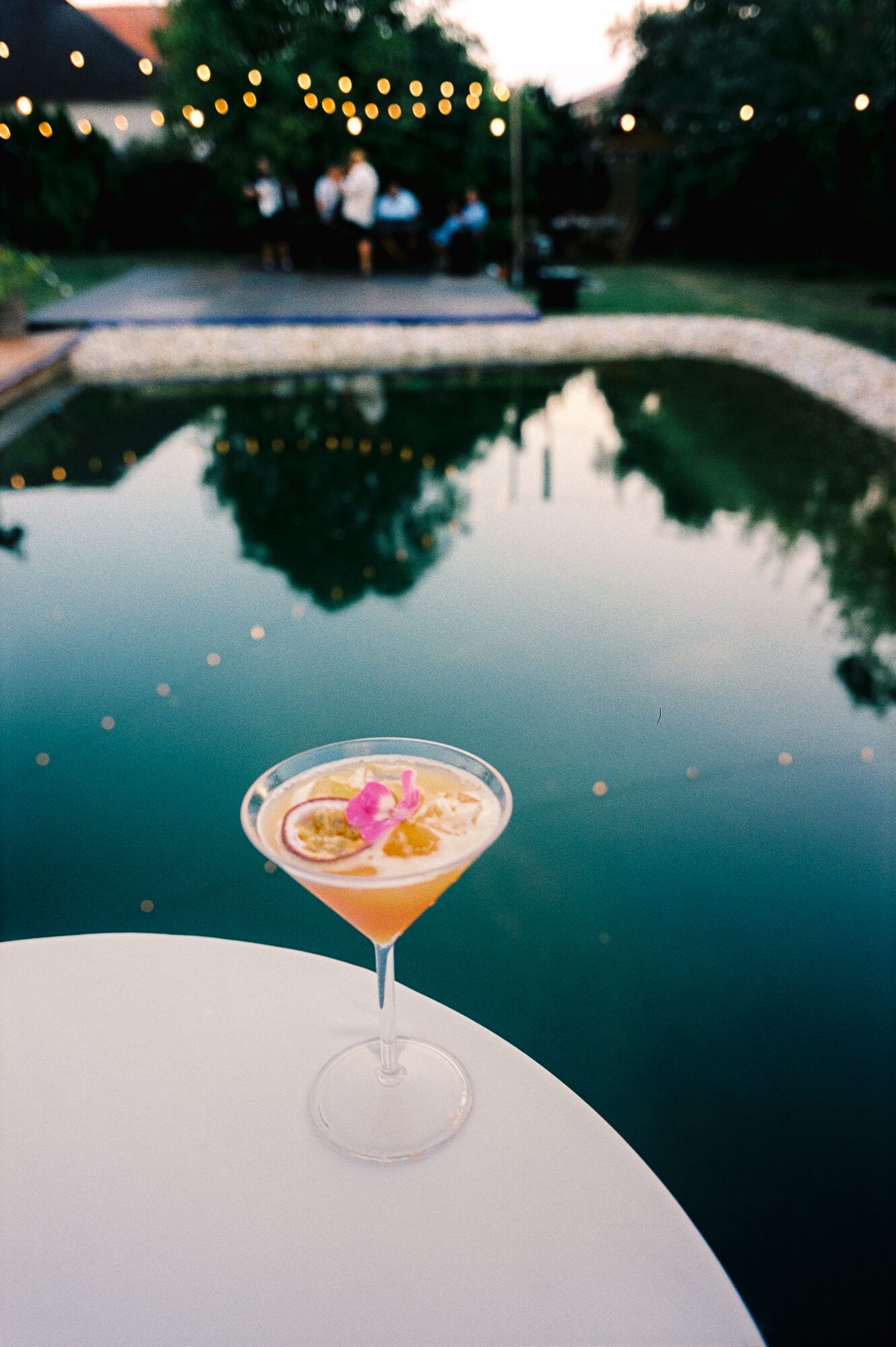 Elegant cocktail with citrus slices and pink flower garnish on white table beside a serene pool at dusk.