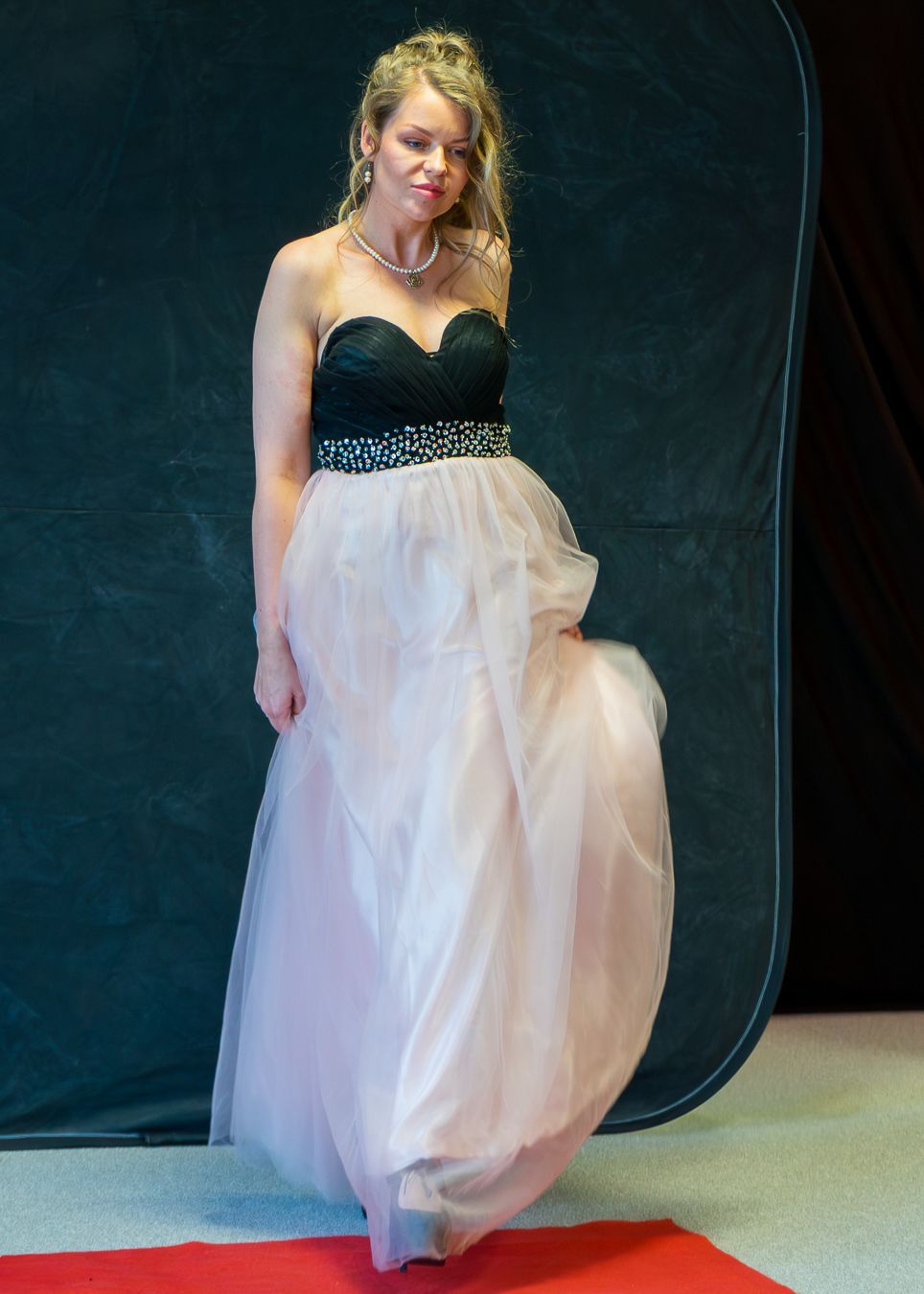 A person in a black and white dress adjusts their gown while standing against a dark backdrop in a studio setting.