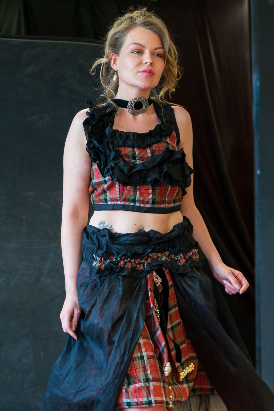 A model wearing a dark ruffled top with plaid skirt details poses against a black backdrop in a fashion photoshoot series.