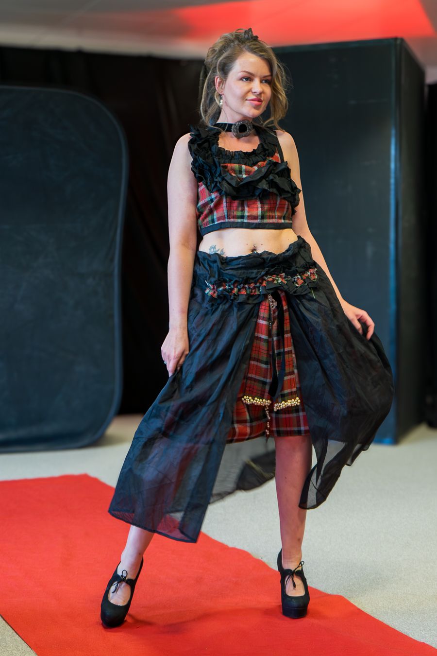 Model wearing a navy blue dress with red plaid scarf poses in dynamic fashion sequence on red and black backdrop.