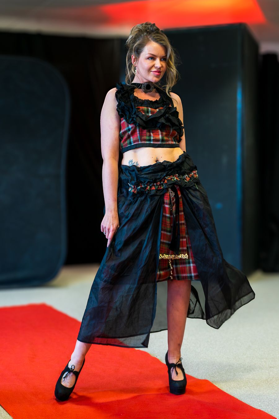Model wearing black babydoll dress with plaid accents strikes dramatic poses on red runway.