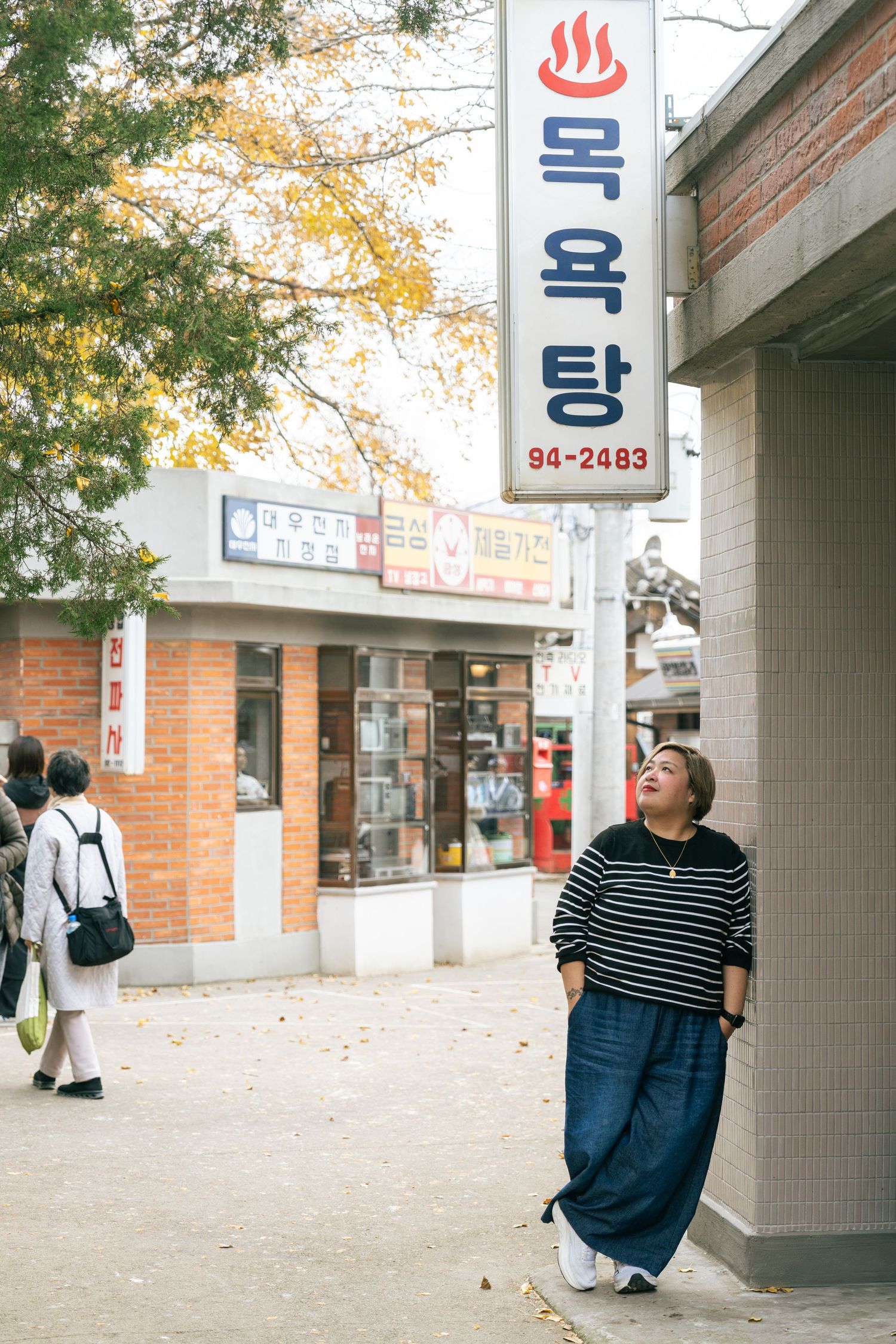 Five connected photos show figures in casual wear standing against a brick wall with Korean signage and autumn leaves.