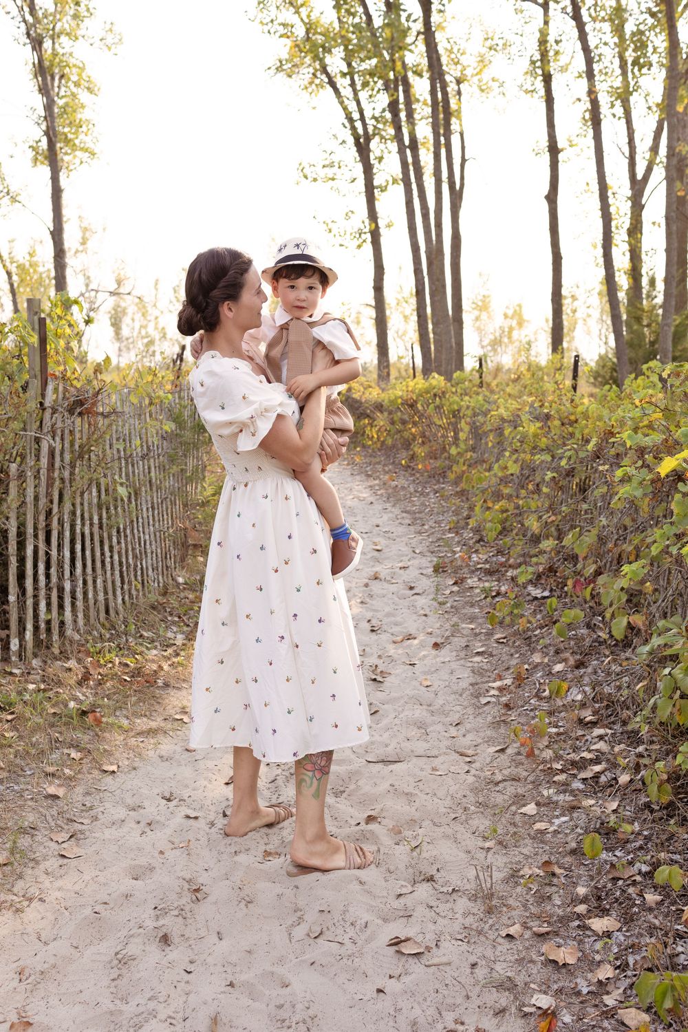 A mother and child walk hand in hand along a sandy wooded path surrounded by autumn foliage.