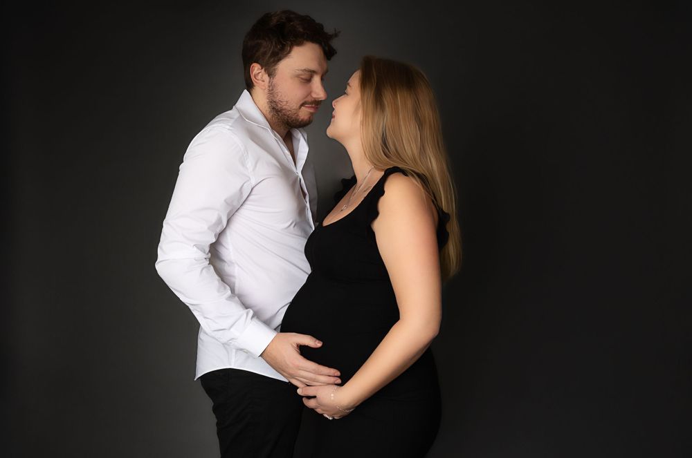Intimate maternity portrait in black dress and white shirt against dark dramatic backdrop.