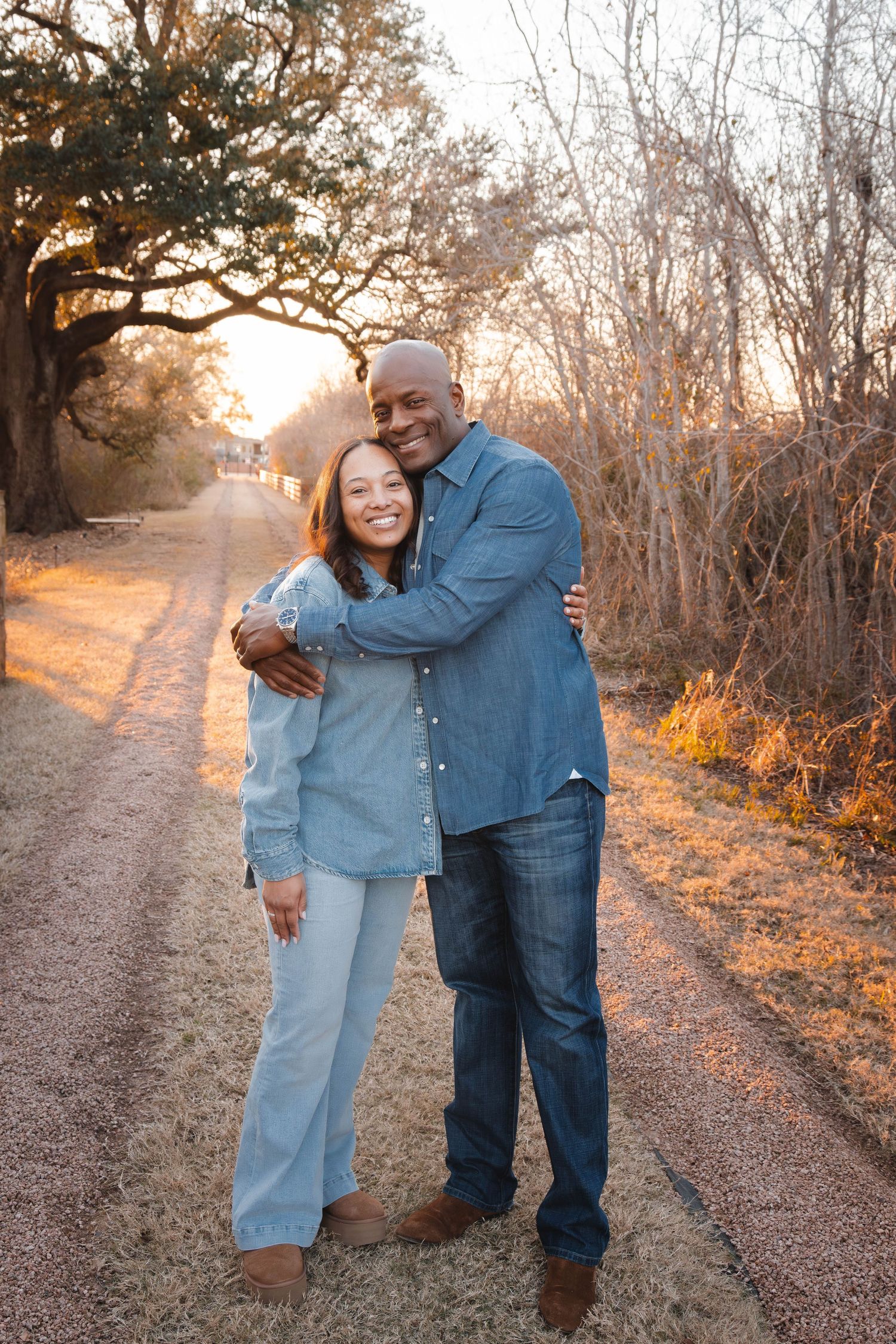 A couple shares an intimate moment on a sunlit autumn path surrounded by bare trees and golden light.